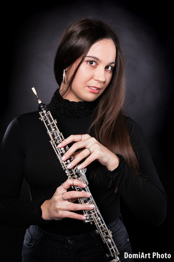 Fuvolát a kezében tartó fiatal hölgy fekete ruhában áll a kamera előtt, fekete háttér előtt. Műtermi fotózás hangszerrel.