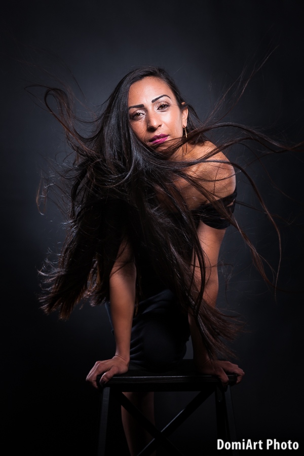 hosszú fekete hajú modell hajol előre és szélgéppel a hajába fújjuk a levegőt, így a hosszú haja szállni kezd. Műtermi fekete hátteres fotózás.
